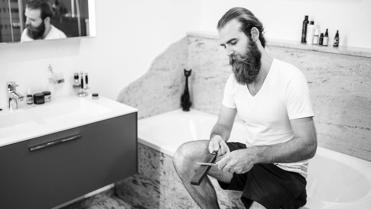 Mann zieht Rasiermesser auf Spannriemen ab auf Badewanne sitzend